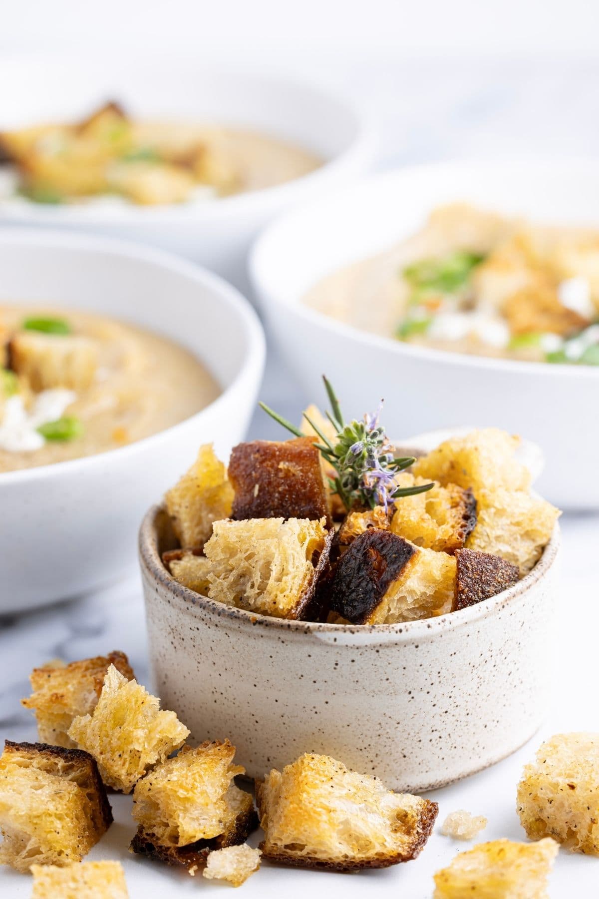 A vertical image of a small speckled dish filled with golden brown sourdough croutons. The croutons are spilling out onto the counter. In the background you can see 3 bowls of vegetable soup, all topped with sourdough croutons and green onions.