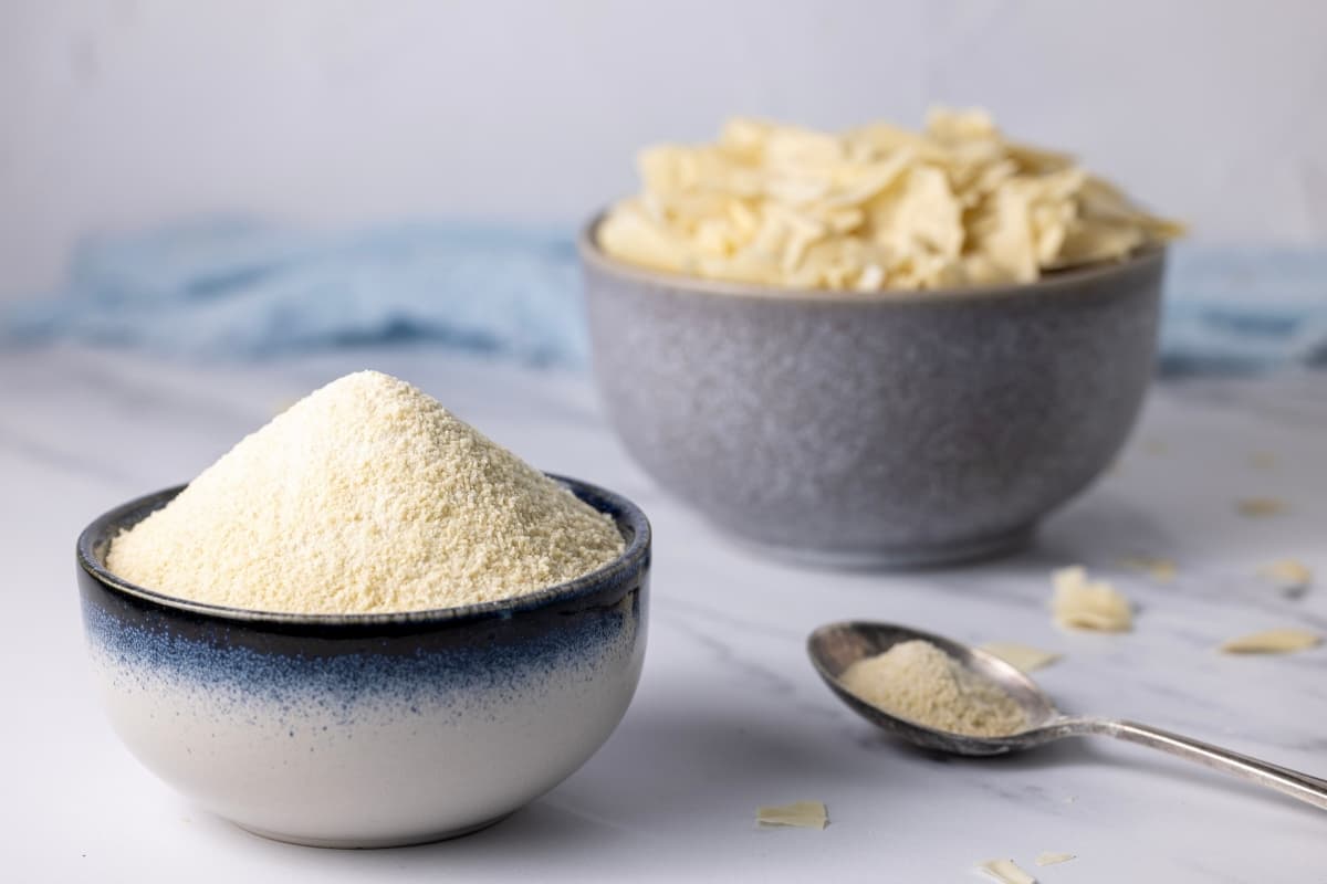 A photo featuring two bowls. The bowl on the right is in the background and is larger. It is full of dried sourdough starter that is still in large chunks and shards. The bowl at the front is smaller and is piled high with dehydrated sourdough starter that has been blitzed into a finer powder. 