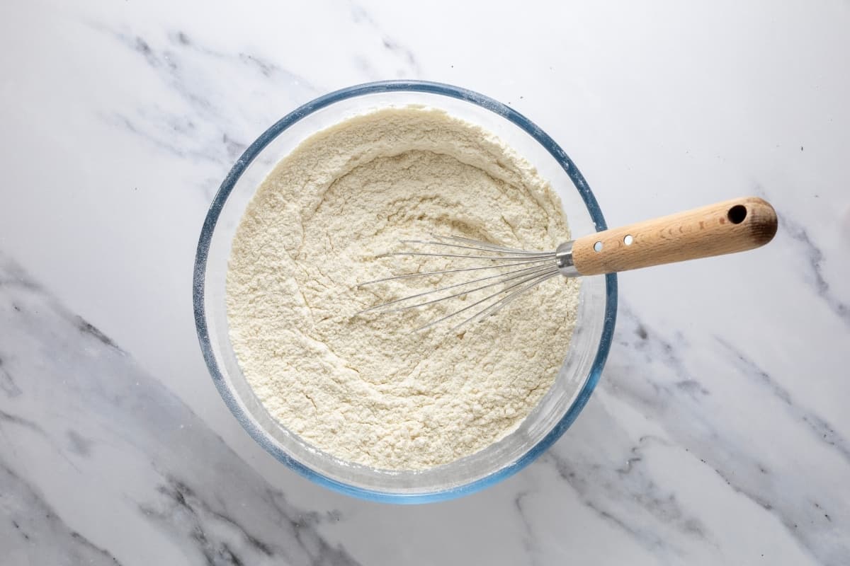 An image of a glass bowl filled with homemade sourdough pancake mix that has been whisked together. The wooden handled whisk is still sitting in the bowl.
