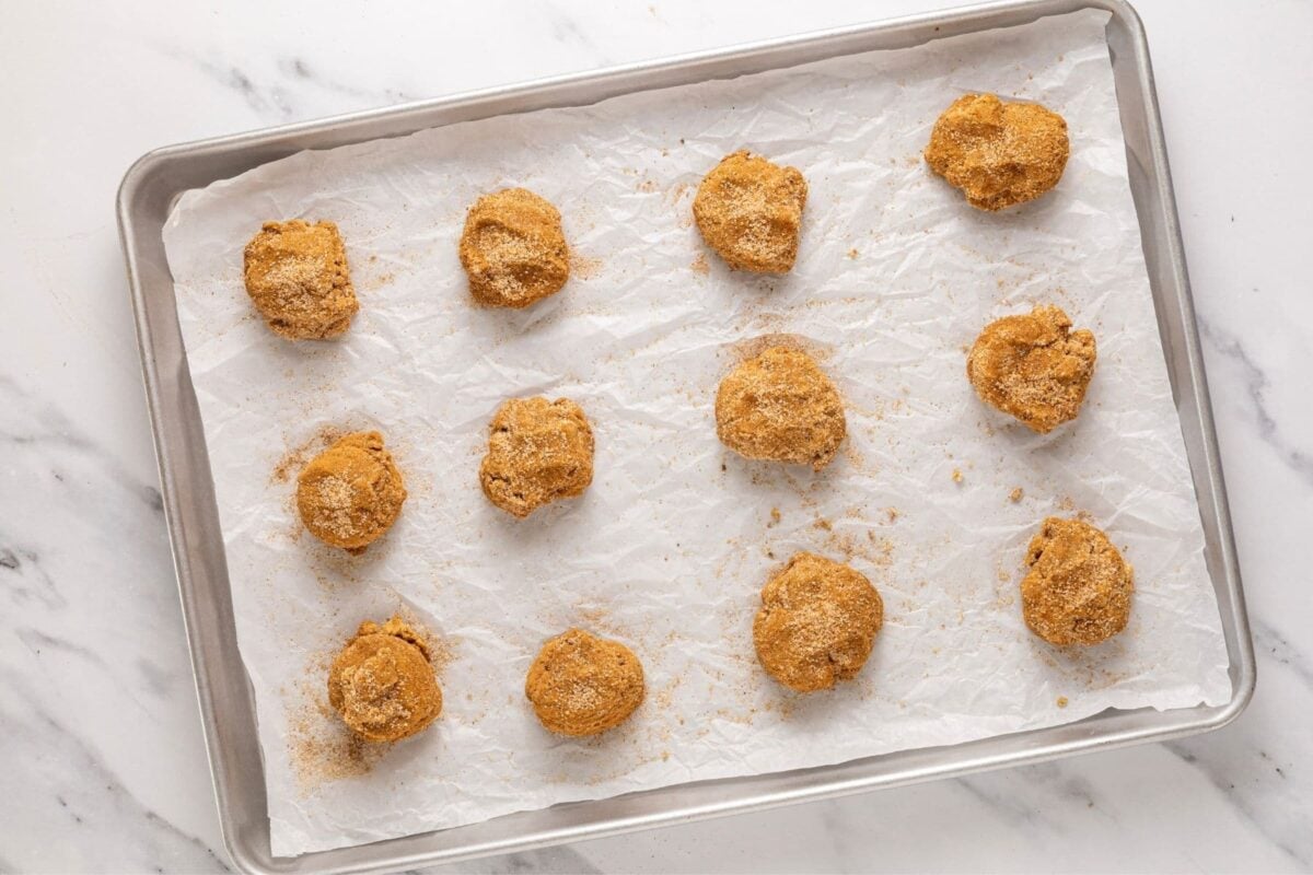 A large baking sheet lined with parchment paper containing 12 sourdough apple cider cookie dough balls that have been pressed down a little.
