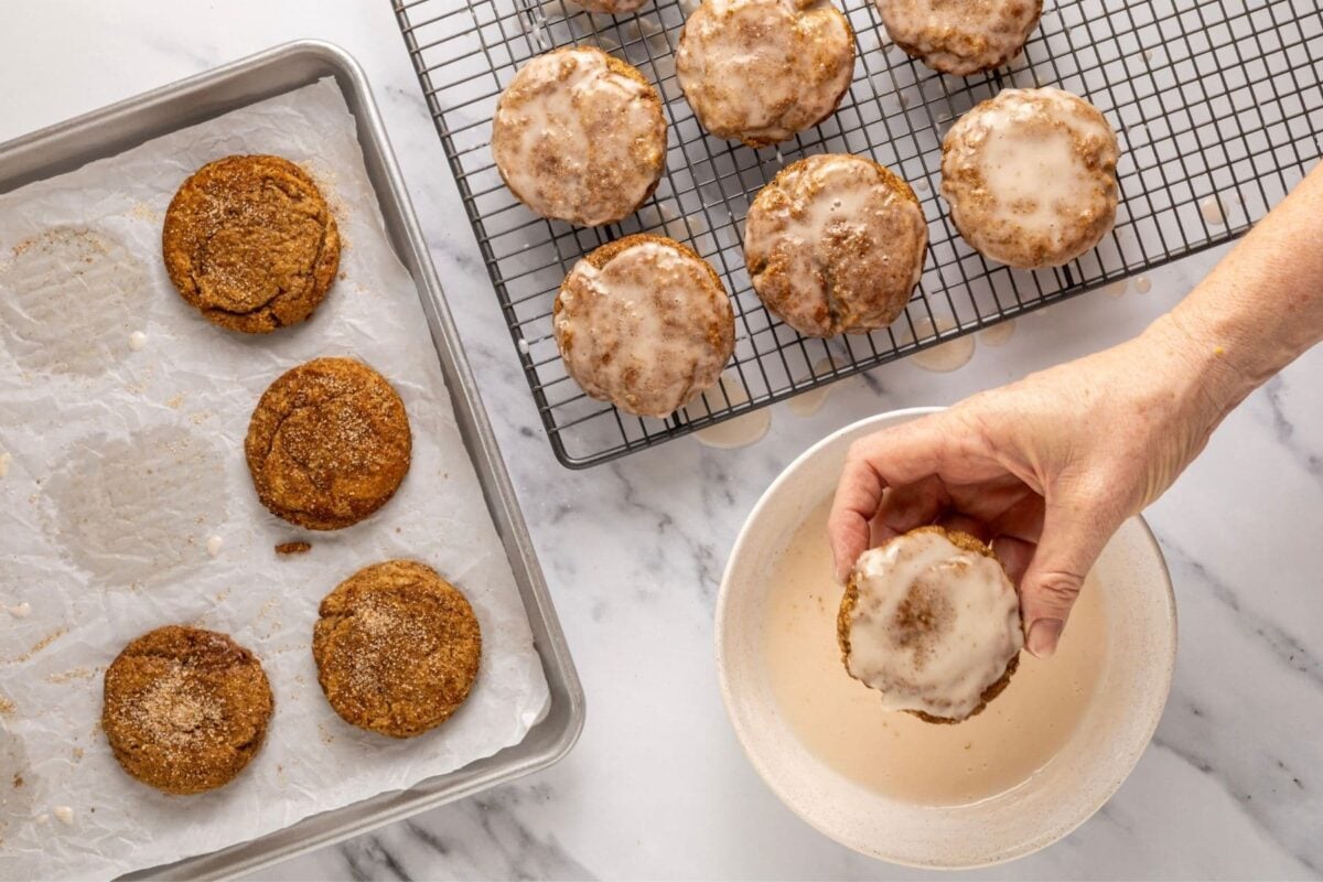 A horizontal image showing how to dip the sourdough apple cider cookies into a small bowl of apple cider glaze. There is hand holding one of the cookies to show how much glaze should be on the the top of each cookie.