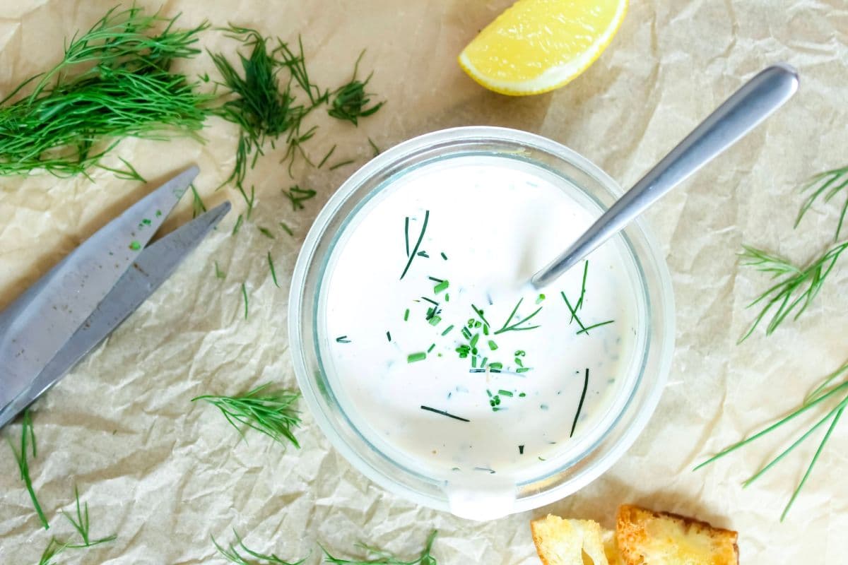 Homemade ranch dressing topped with fresh dill and chives, on a piece of parchment paper next to lemon and croutons.
