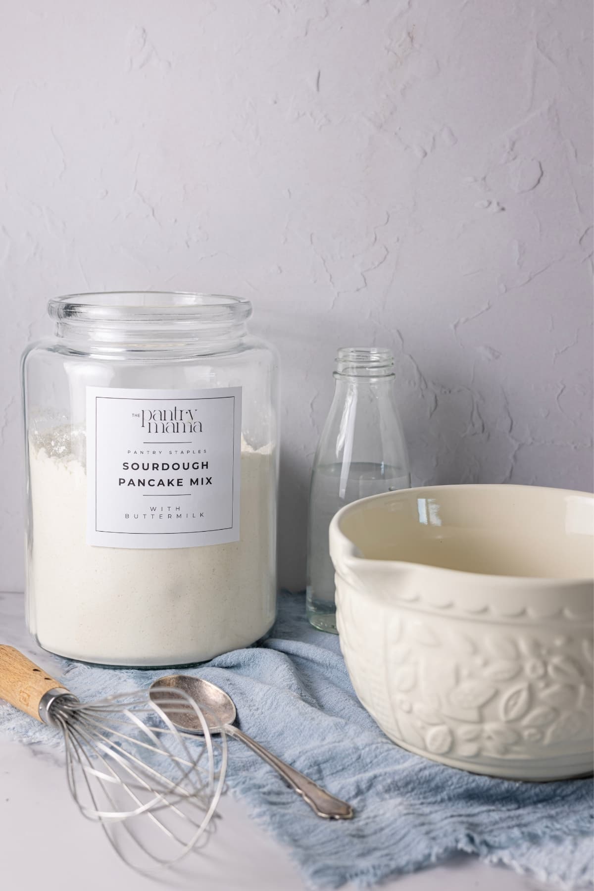 A vertical image showing a large glass jar of homemade sourdough pancake mix sitting a blue linen napkin with a vintage spoon and wooden handled whisk. There is a cream embossed batter jug sitting to the right of the photo.