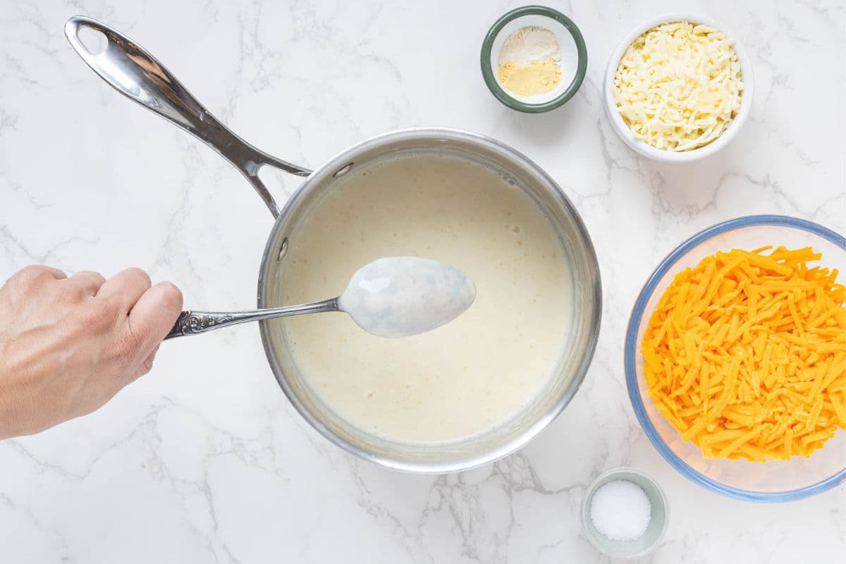 An image showing a spoon held over a saucepan of white sauce. The sauce is coating the back of the spoon. Next to the pot there is a bowl of shredded cheddar cheese, parmesan cheese and garlic and mustard powder.