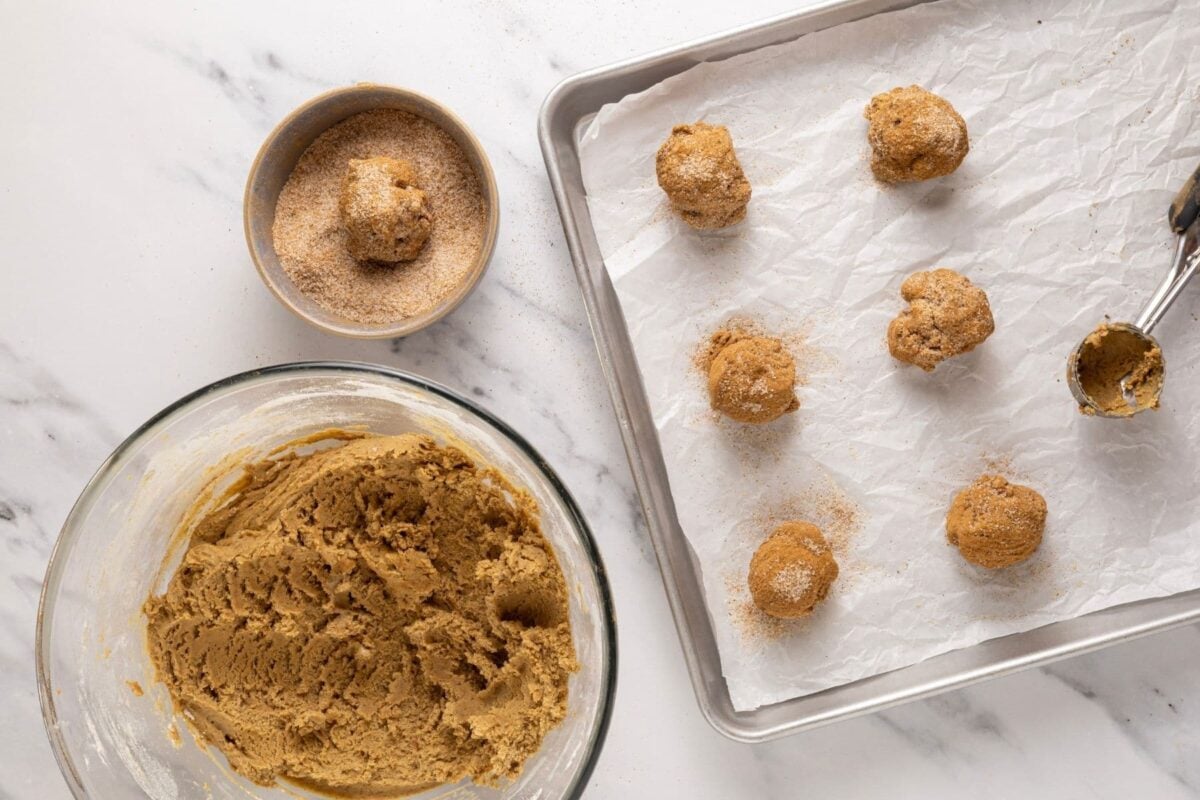 A horizontal image of a bowl of sourdough apple cider cookie dough sitting along side a small bowl of spiced sugar. There is a baking tray with cookie dough balls on it. The photo is demonstrating the process of scooping the cookie dough balls into the spiced sugar before placing them onto the tray.