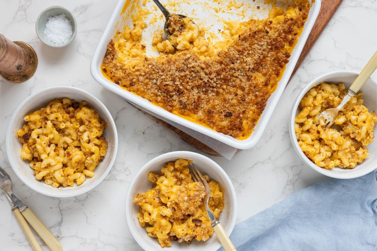A horizontal image showing a large white dish filled with sourdough mac and cheese in the top centre of the image. There are three bowls filled with servings of the mac and cheese