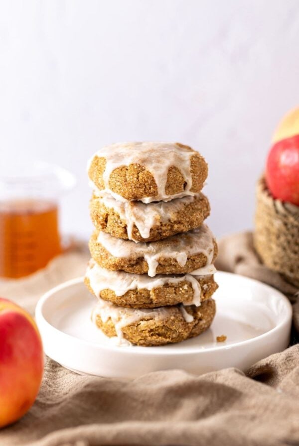A stack of 5 sourdough apple cider cookies that have been drizzled with glaze and placed onto a white plate. You can see a beaker of apple cider in the background of the photo, as well as a basket of apples to the right of the cookies.