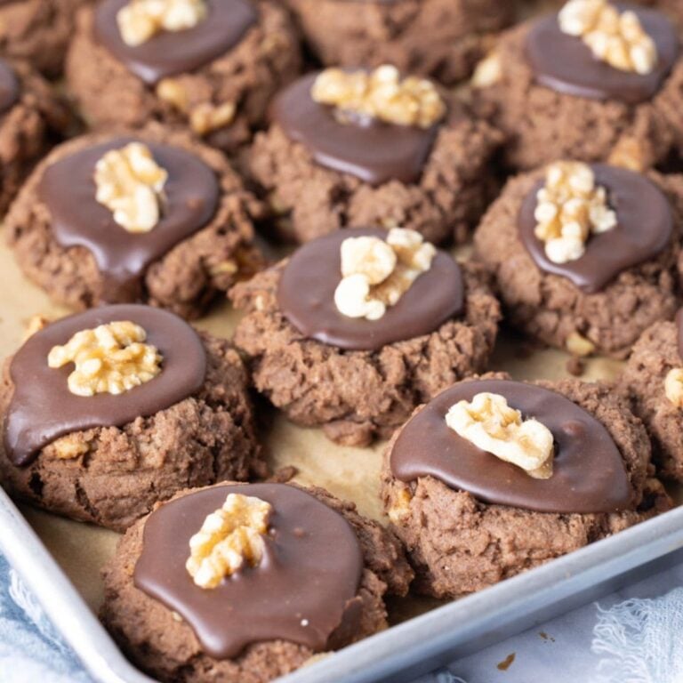 Close up of frosted sourdough chocolate walnut cookies with a walnut half on top on a baking sheet.