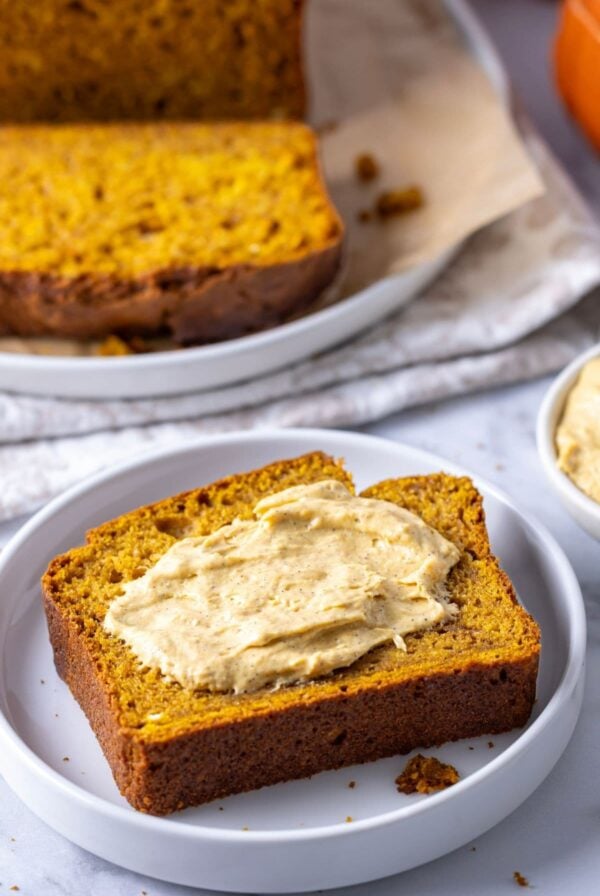 Vertical image of a slice of sourdough discard pumpkin loaf that has been placed on a white plate and spread with spiced pumpkin cream cheese.