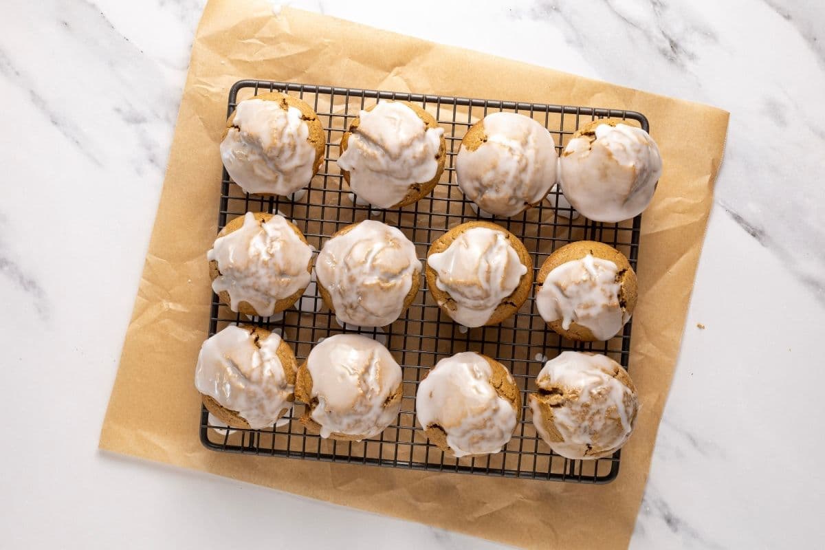 A batch of 12 sourdough gingerbread muffins drizzled in white sugar glaze. The muffins are sitting on a wire cooling rack with a piece of parchment paper underneath to catch the drips of glaze. The photo has been taken top down.