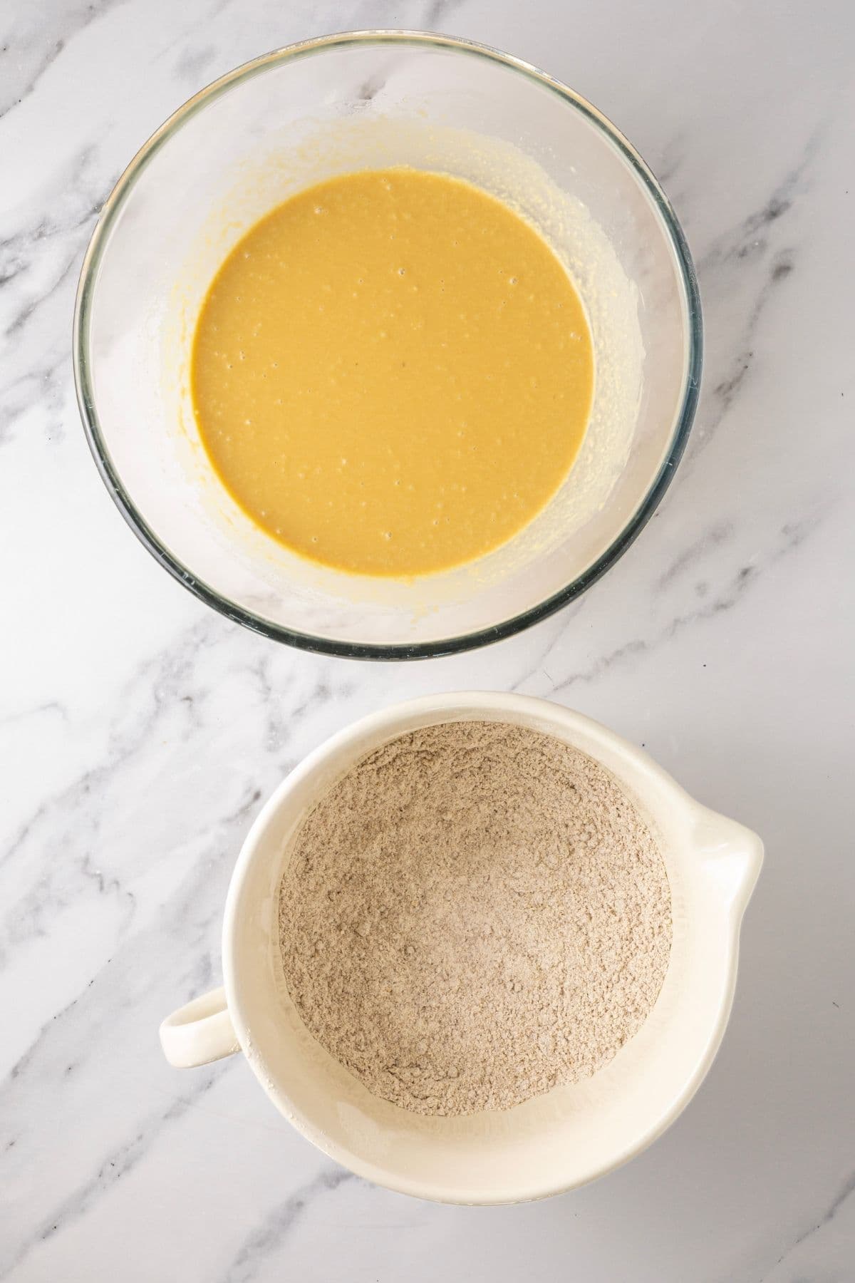 An image showing two bowls. The top bowl is glass and filled with liquid ingredients and the bowl on the bottom is ceramic and is filled with dry ingredients.