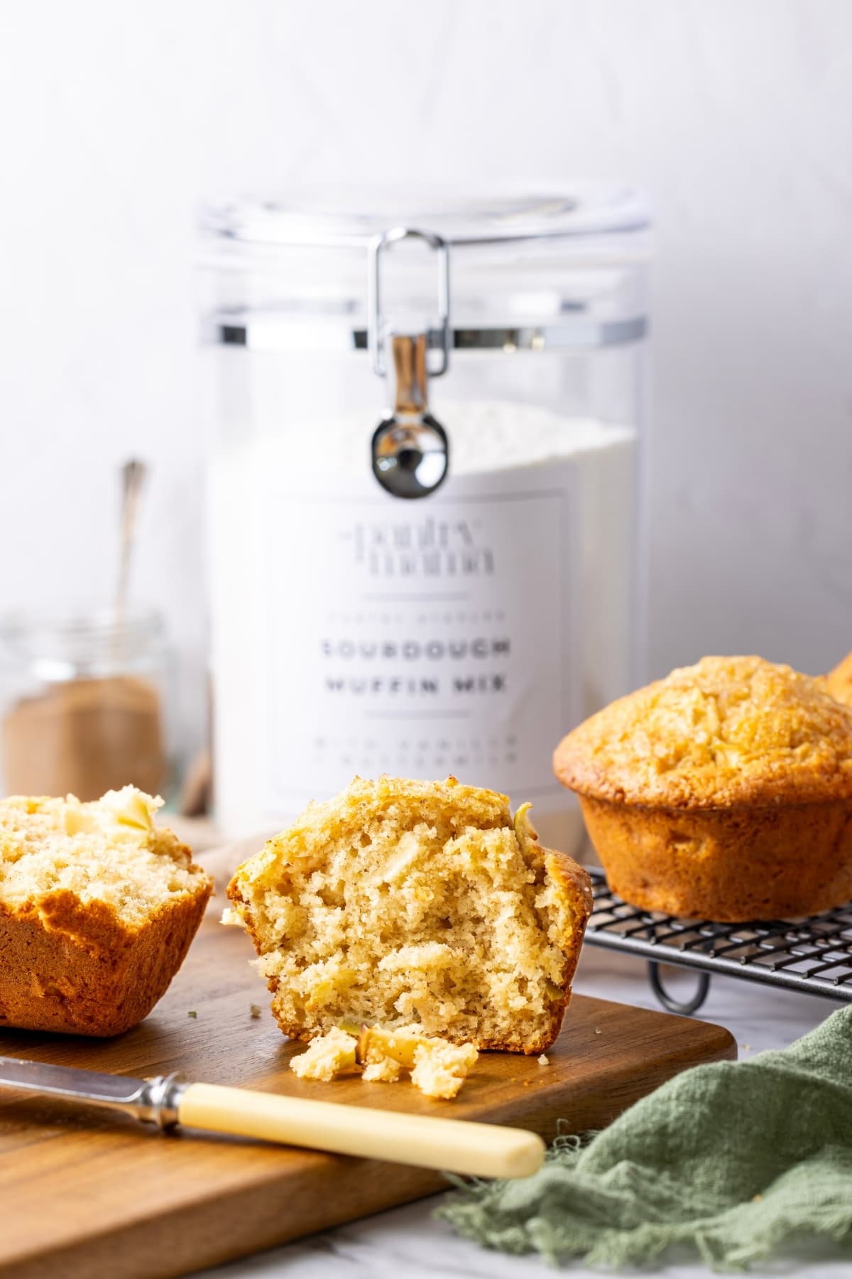 A vertical image showing a jar of sourdough muffin mix in the background. In the foreground there are fresh muffins that have been made using the dry mix. The one at the very focal point of the image has been sliced open.