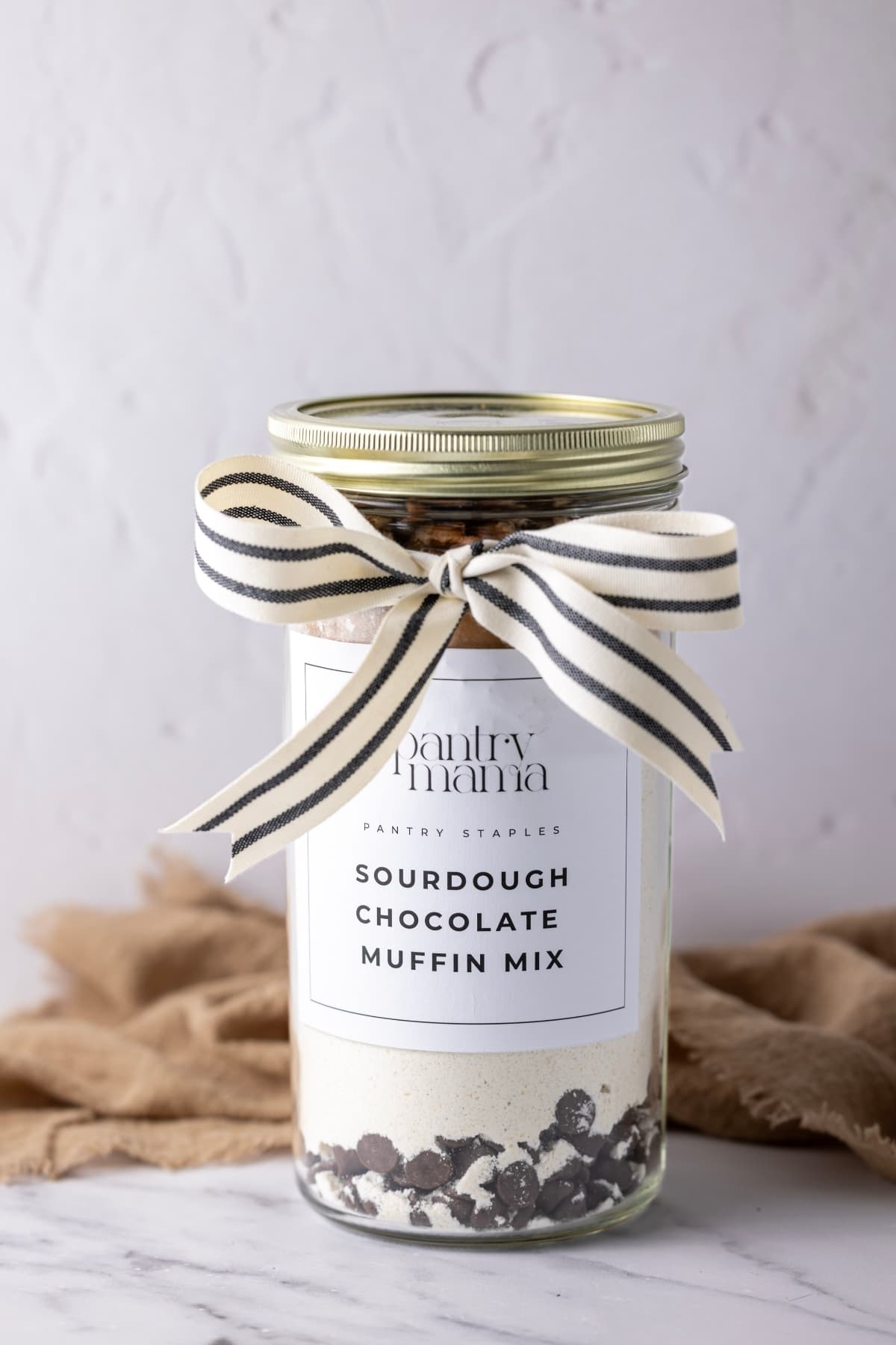A tall jar with a gold lid filled with layers of dry sourdough muffin mix and chocolate chips. It has a cream and black ribbon tied at the top and a black and white label on the jar which reads "Chocolate Sourdough Muffin Mix".