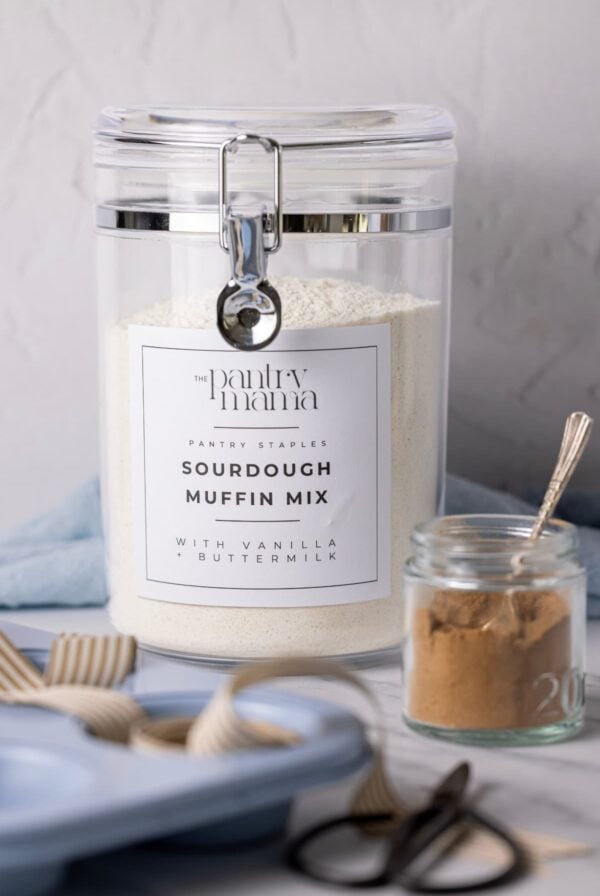 A large clear plastic container with a flip top lid containing dry sourdough muffin mix. It has a black and white label on the front labelled "sourdough muffin mix". There is a small jar of cinnamon in front of the canister and a blue muffin pan at the front of it.