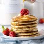 An image showing a stack of sourdough pancakes topped with bright red strawberries. There is a blue linen napkin sitting under the plate of pancakes. There is a large glass jar of homemade sourdough pancake mix in the background of the photo.