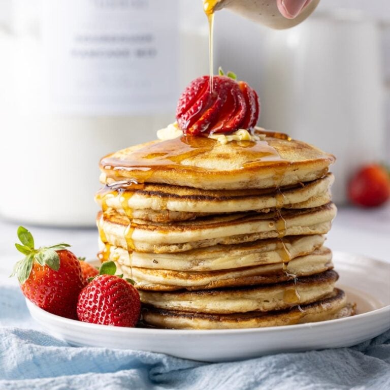 An image showing a stack of sourdough pancakes topped with bright red strawberries. There is a blue linen napkin sitting under the plate of pancakes. There is a large glass jar of homemade sourdough pancake mix in the background of the photo.