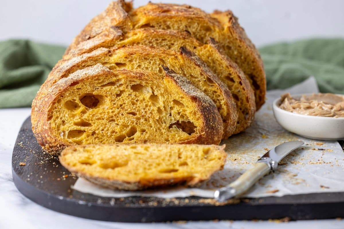 An artisan pumpkin sourdough loaf that has been sliced and arranged so that you can see the cut face of the slices - the golden, tender crumb and crisp crust of the loaf. There is a green linen napkin draped in the background of the image.