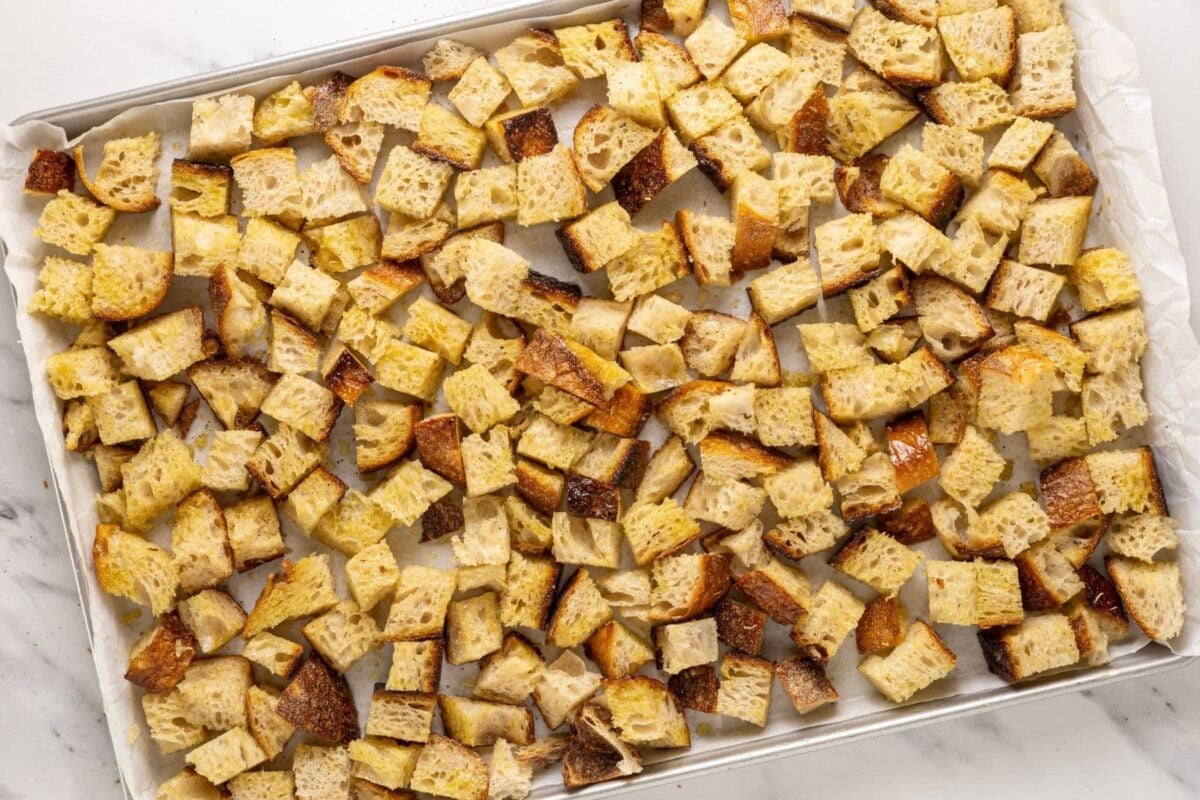 A large baking tray filled with a single layer of cubed sourdough bread that has been tossed in olive oil and seasoning. These sourdough croutons are unbaked.