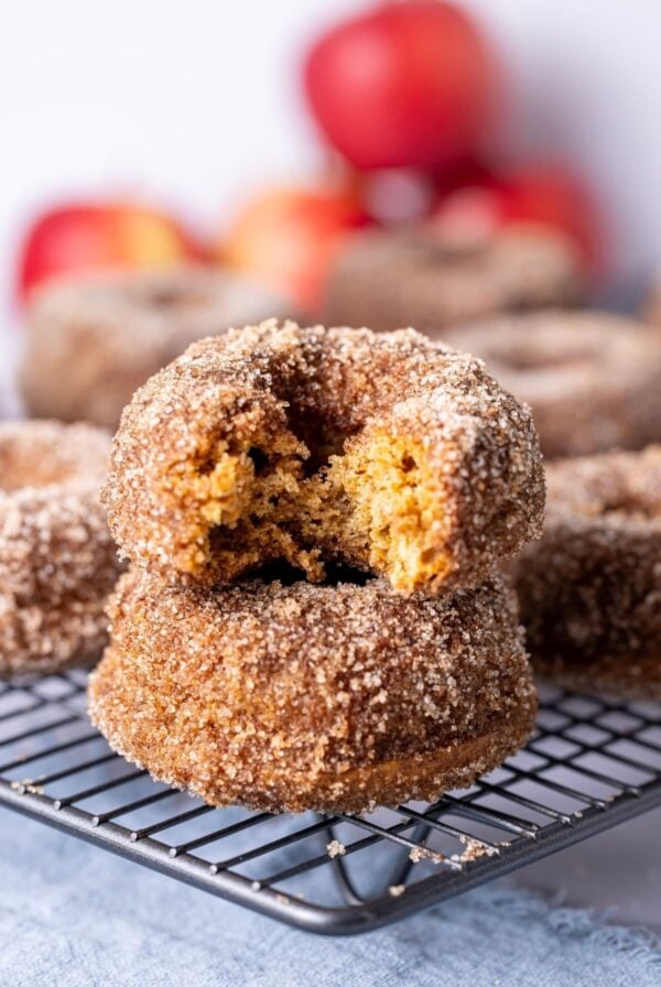 Two sourdough apple cider donuts stacked on top of each other. The donut on the top has a bite taken out of it. There are some red apple in the background of the photo.