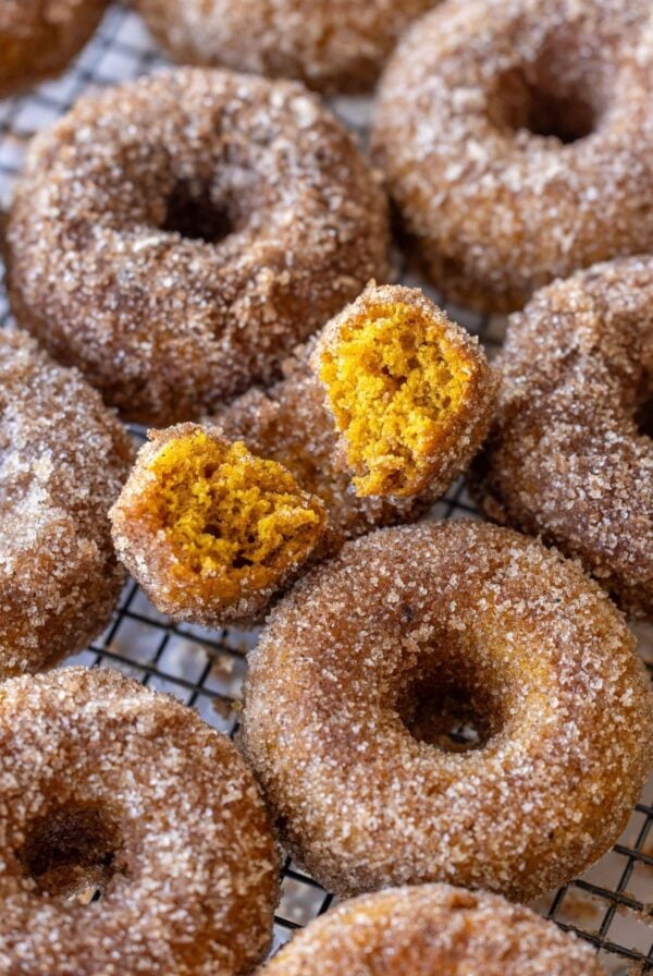 An image taken from above of baked sourdough sugared donuts layed out on a wire cooling rack. The donut in the middle has a bite taken out of it so you can see the gorgeous orange color and texture inside.