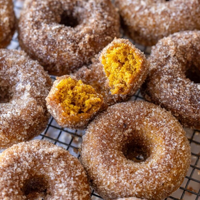 An image taken from above of baked sourdough sugared donuts layed out on a wire cooling rack. The donut in the middle has a bite taken out of it so you can see the gorgeous orange color and texture inside.