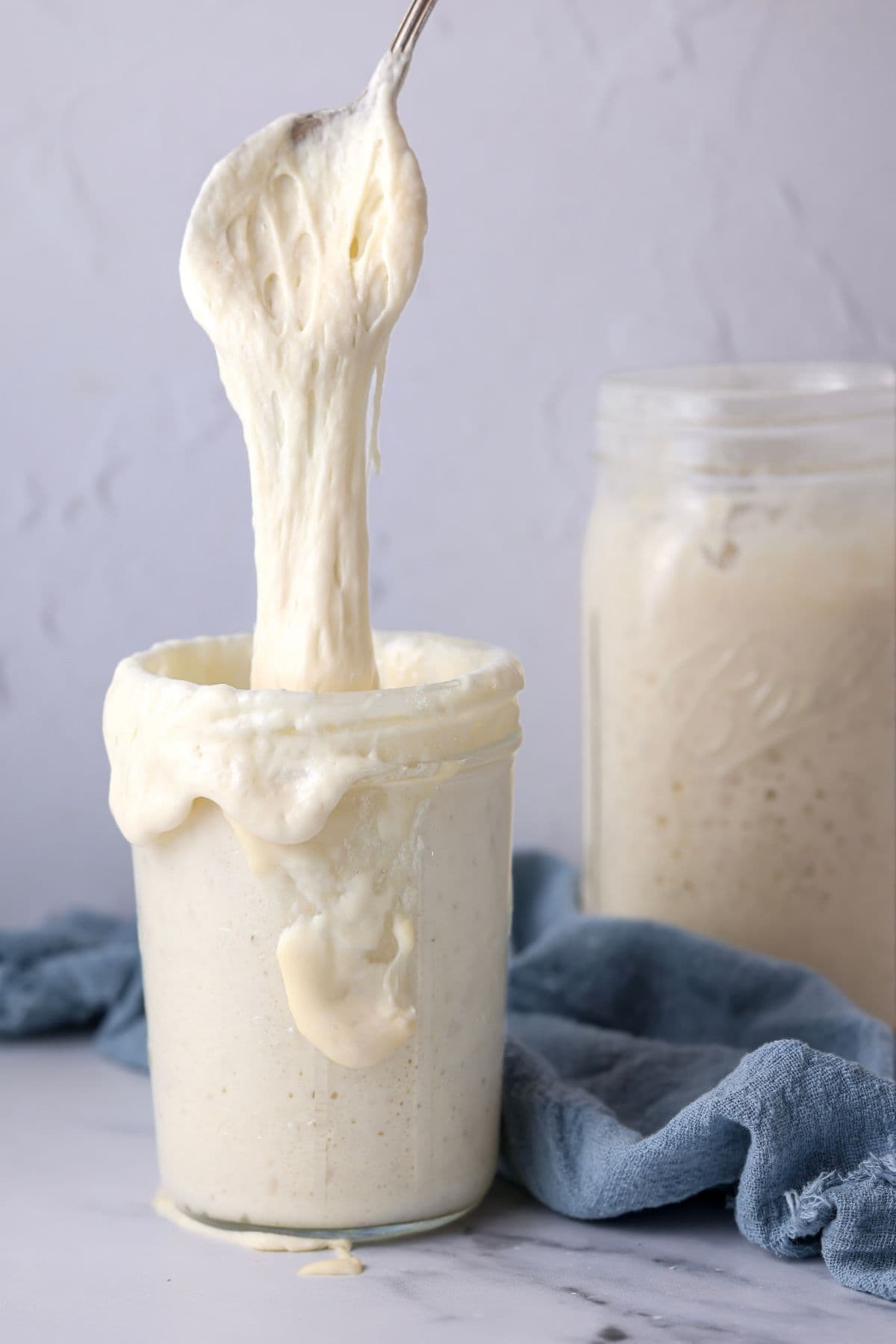 A jar of sourdough starter that has overflowed and is sliding down the side of the jar. There is a spoon that is pulling some of the sourdough starter out of the jar.