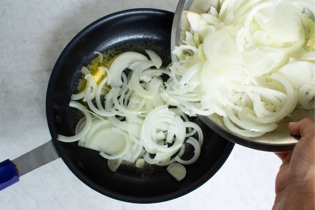 Thinly sliced onions being added to a skillet with melted butter to make caramelized French onion dip.