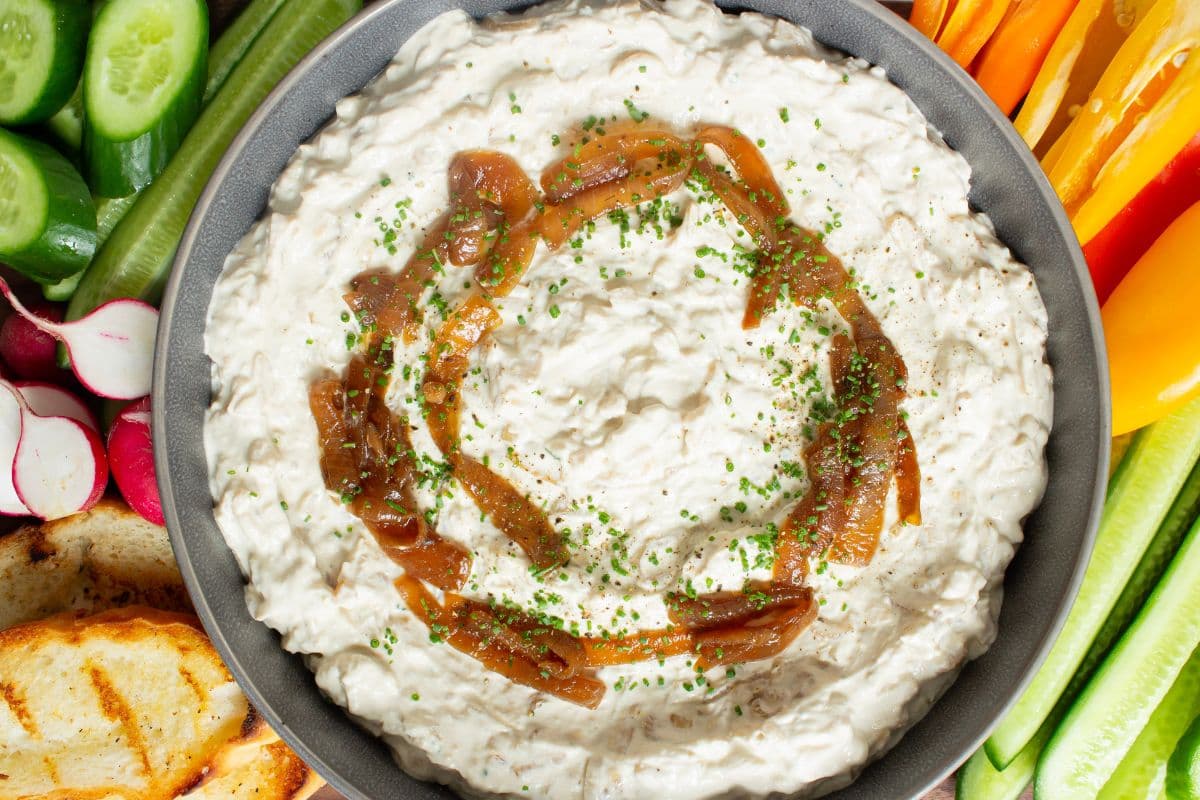 Overhead view of caramelized French onion dip topped with caramelized onions and fresh chives, surrounded by cucumber, radishes, peppers, and toasted sourdough bread.