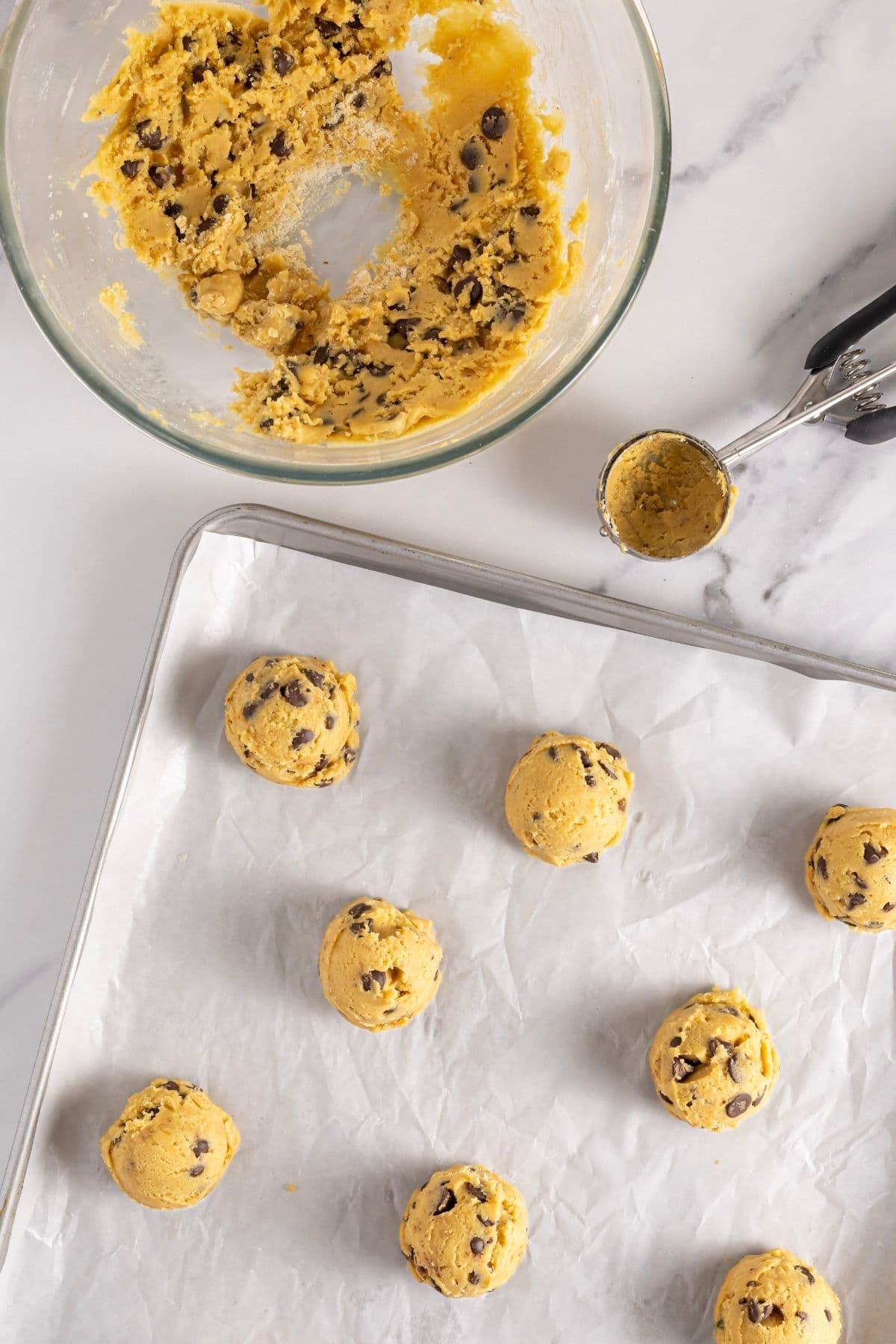 A glass bowl filled with sourdough chocolate chip cookie dough. There are some cookie dough balls that have been scooped out onto a parchment-lined baking tray.