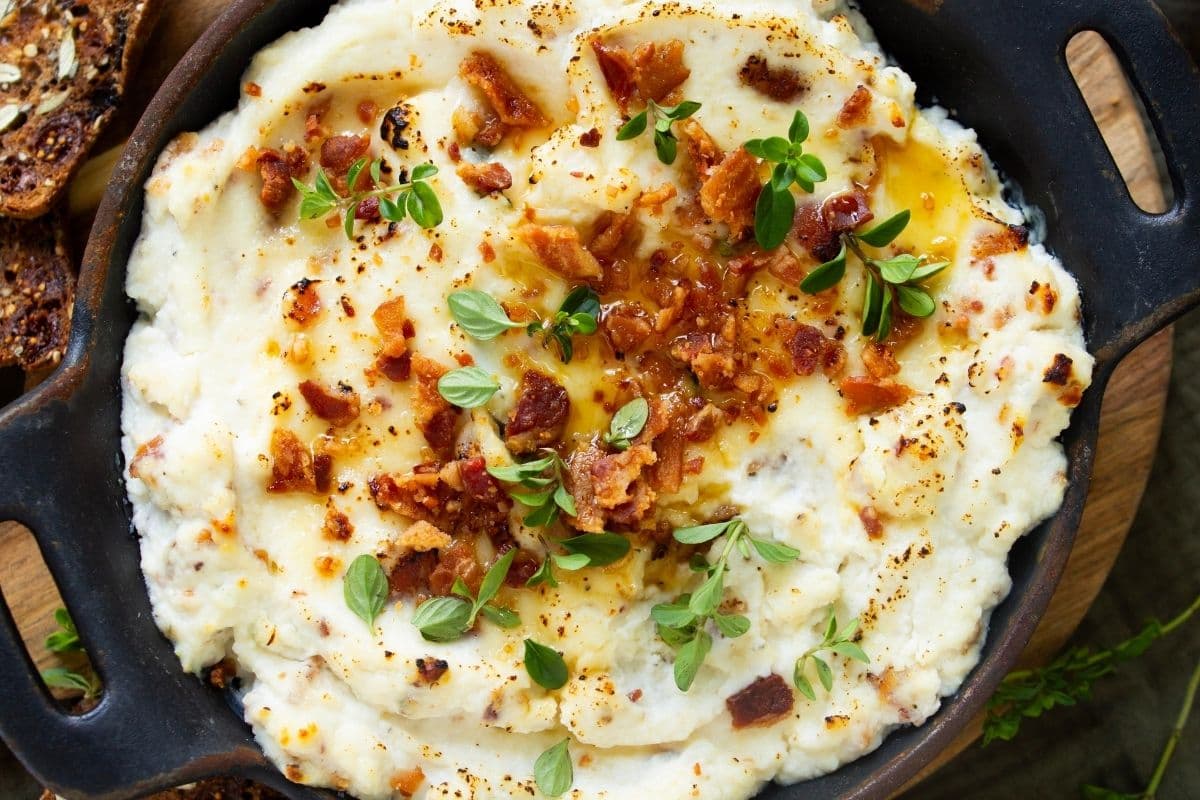 A horizontal image of maple bacon baked ricotta dip topped with bacon, green onions and a drizzle of maple syrup. The dip is being served in a round cast iron dish.