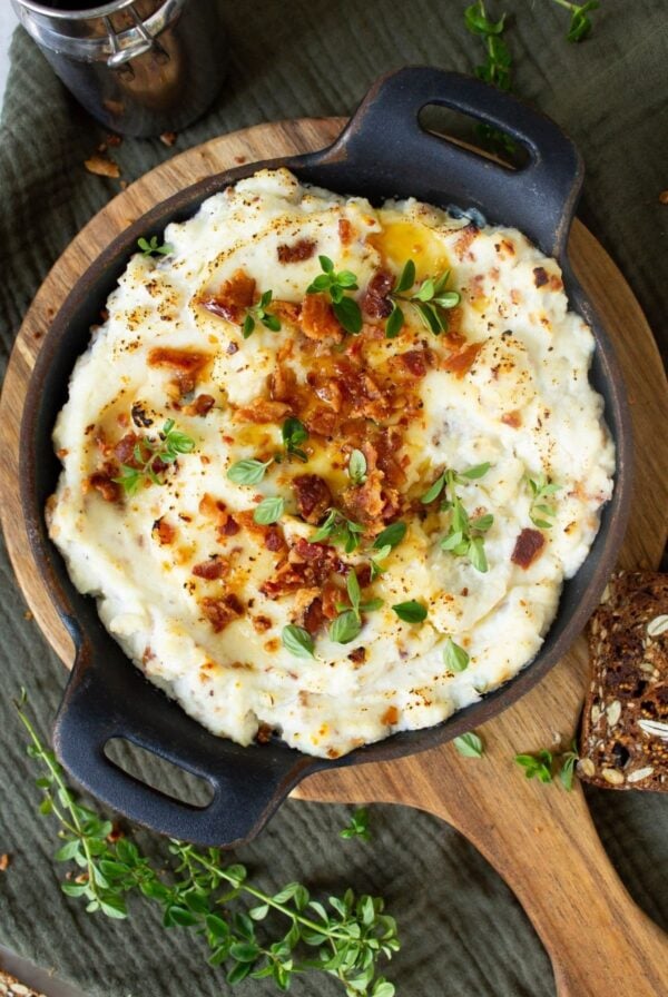 A horizontal image of maple bacon baked ricotta dip topped with bacon, green onions and a drizzle of maple syrup. The dip is being served in a round cast iron dish sitting on a round wooden board. There is some fresh thyme scattered around the image.
