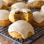 A close up image of a batch of soft sourdough pumpkin spice cookies that have been drizzled with pumpkin glaze sitting on a wire cooling rack. The cookie in the centre of the photo has a bite taken out so you can see the texture inside.