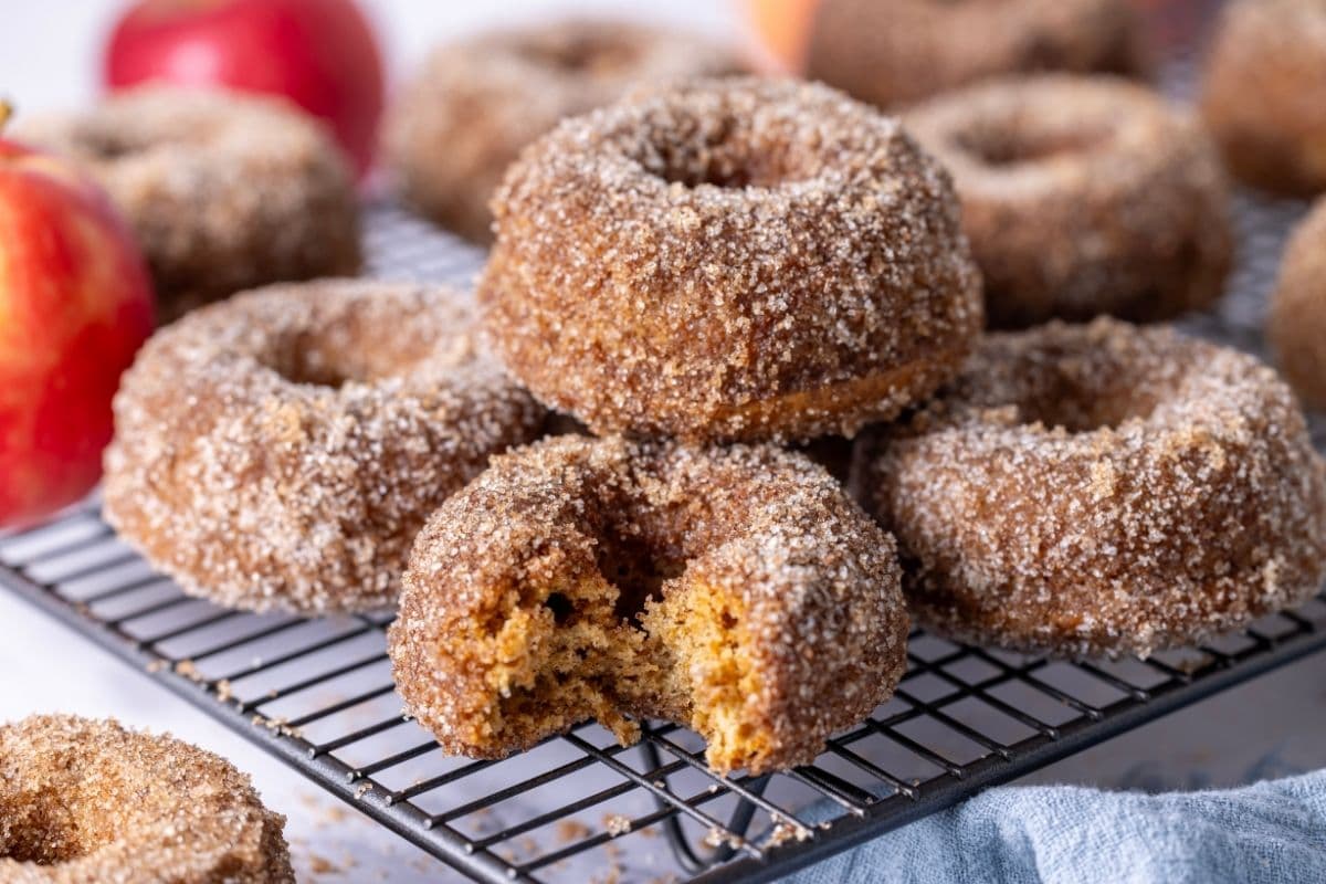 A horizontal image showing a batch of sourdough apple cider donuts cooling on a wire rack. There is a bite taken out of one of the donuts showing the texture inside.