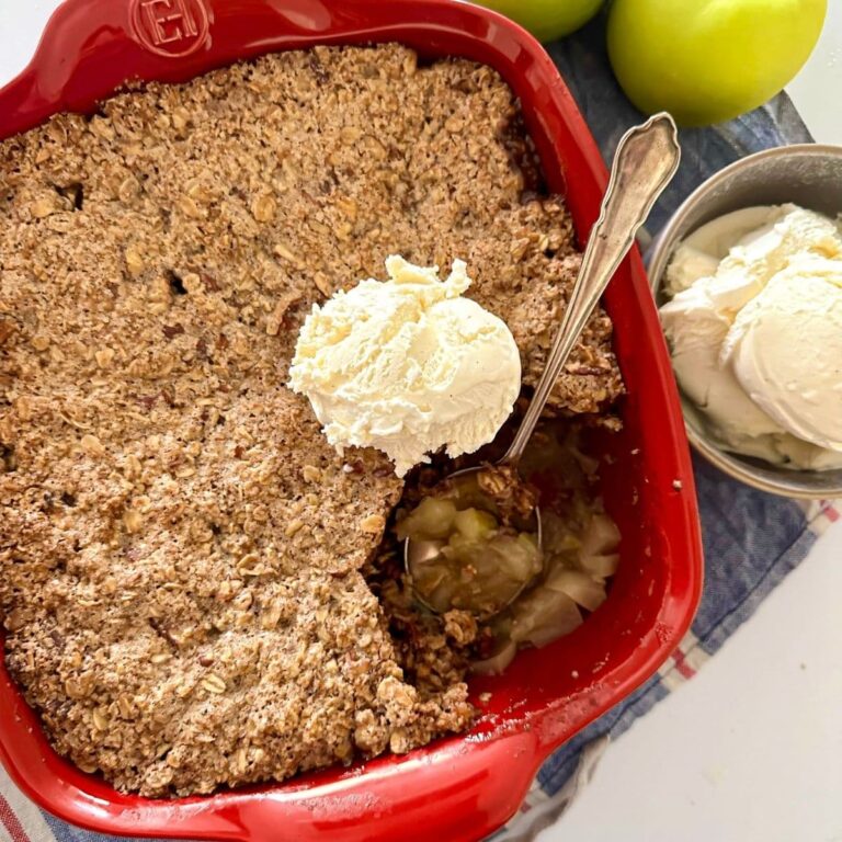 A red square baking dish with a freshly baked sourdough apple crisp topped with vanilla ice cream. A scoop has been taken out so you can see the sweet apple filling underneath.