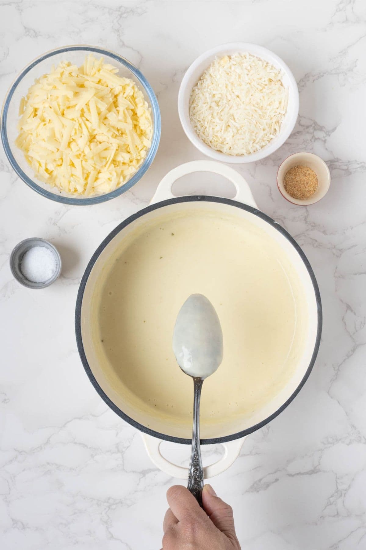 Smooth, creamy cheese sauce in a saucepan with shredded cheese, Parmesan, and seasonings ready to be added with a hand holding a spoon to show roux thickness.