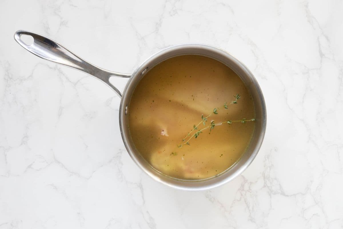 A stainless steel pot filled with chicken breasts, thyme and half the chicken stock.