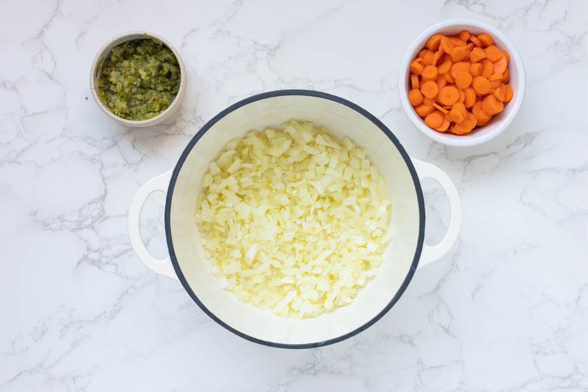A heavy based cream pot with translucent onion and garlic that have been softened. There is a also a small bowl of celery and carrot next to the pot.