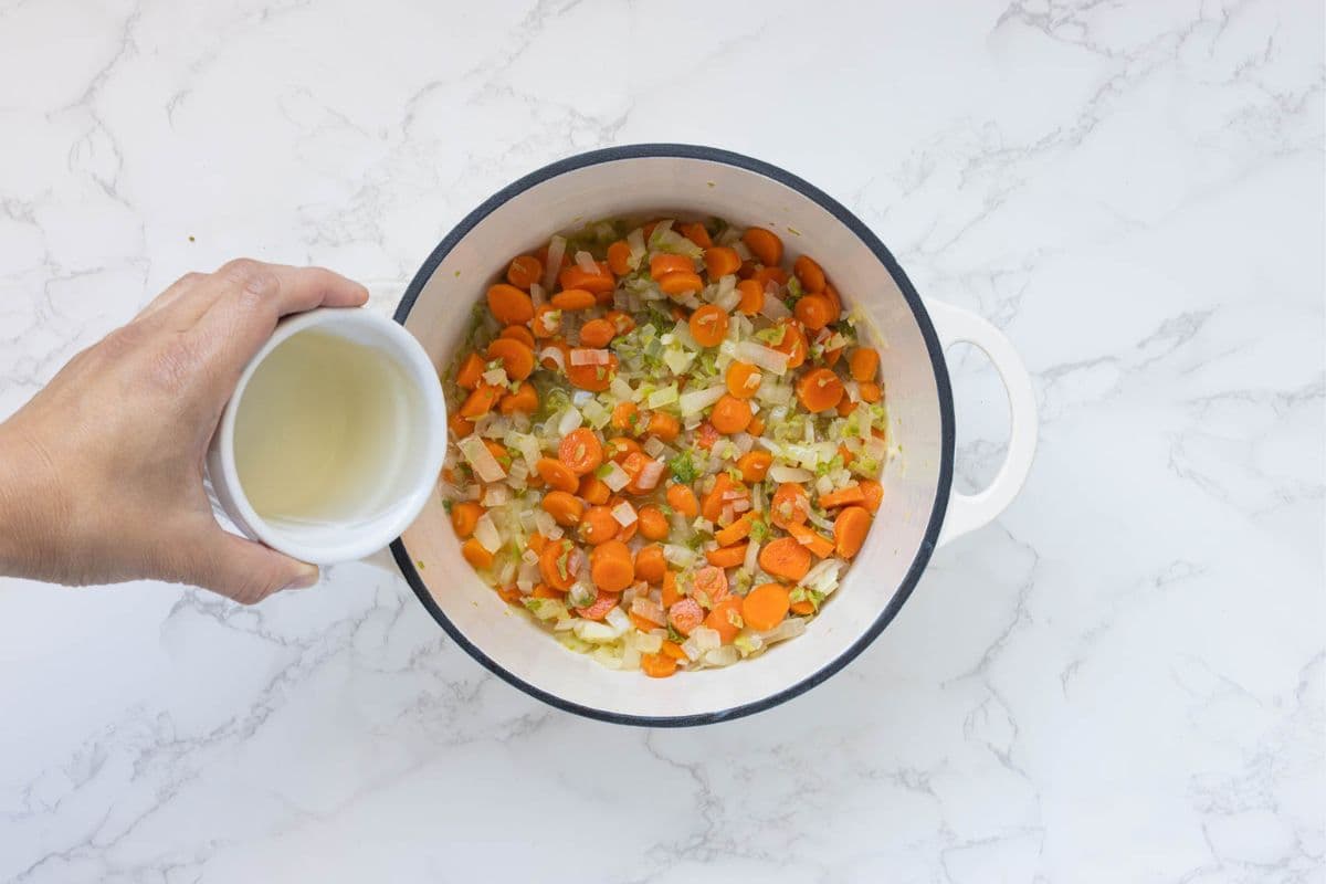 A large cream pot with all the softened vegetables in the bottom. There is a hand tipping a small bowl of white wine into  the pot.