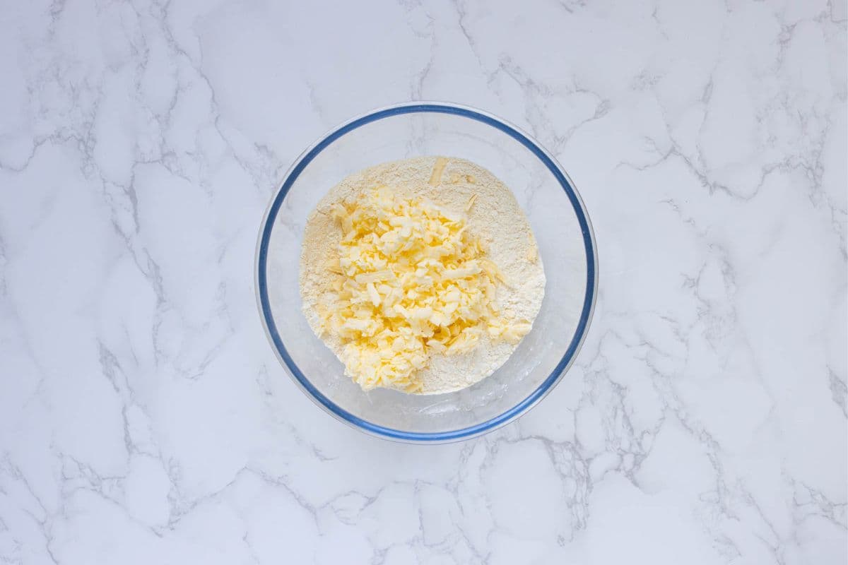A glass bowl filled with the dry ingredients topped with cold, grated butter.