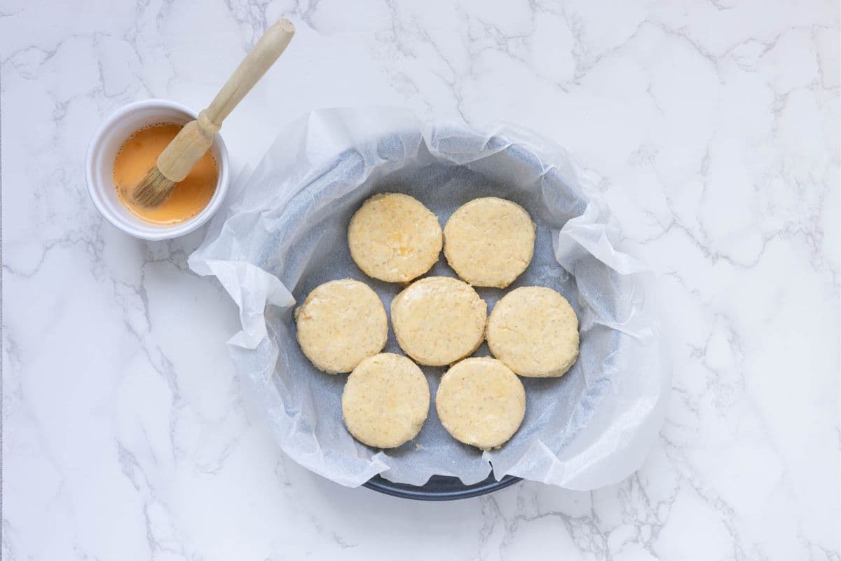 A round cake pan lined with parchment paper and filled with 7 sourdough cornbread biscuits that have been brushed with an egg wash.