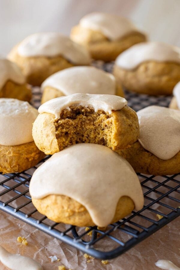 A close up image of a batch of soft sourdough pumpkin spice cookies that have been drizzled with pumpkin glaze sitting on a wire cooling rack. The cookie in the centre of the photo has a bite taken out so you can see the texture inside.