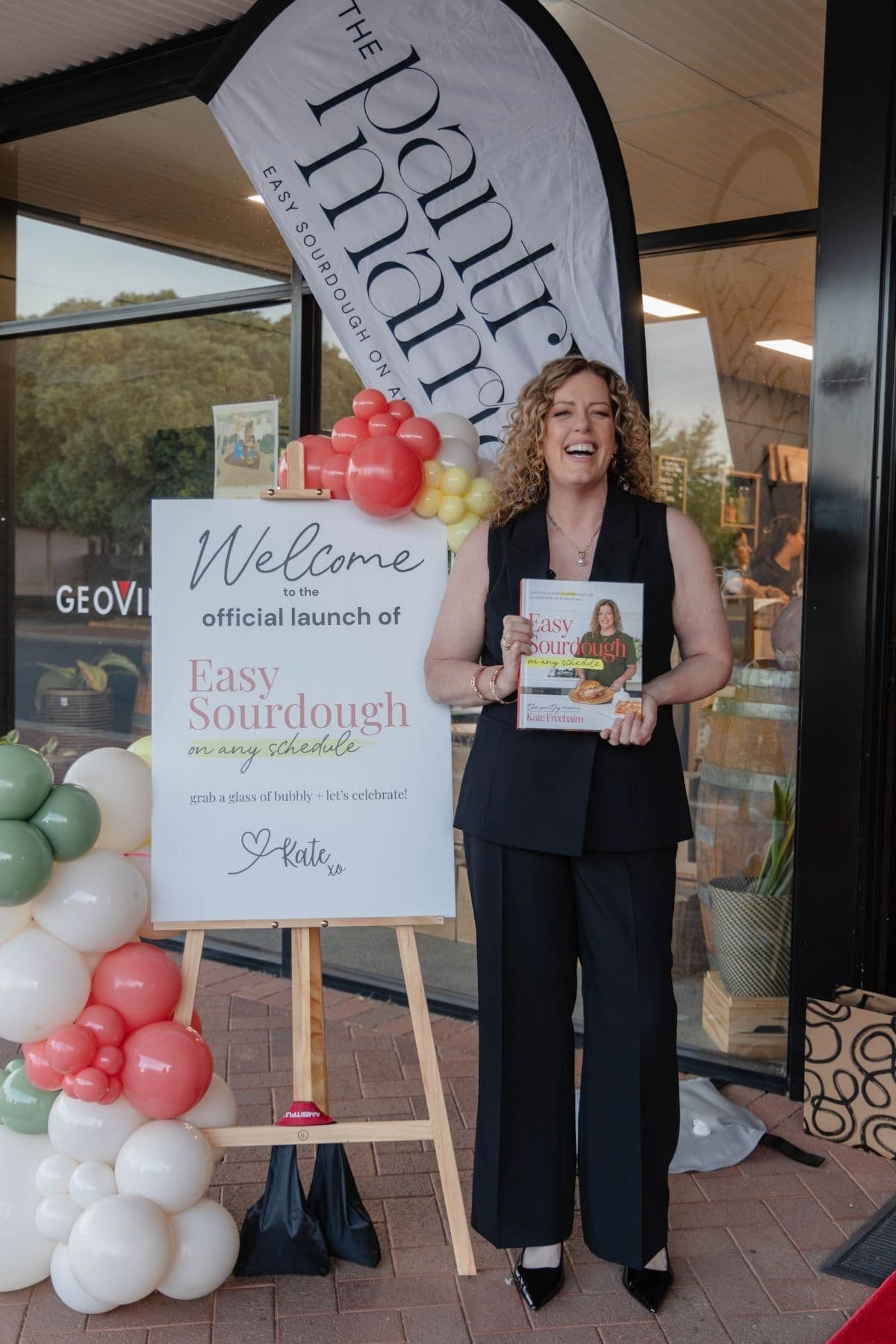Kate Freebairn from The Pantry Mama wearing a black suit and holding a copy of her book Easy Sourdough on Any Schedule next to a sign that says "Welcome to the Official Launch of Easy Sourdough On Any Schedule".