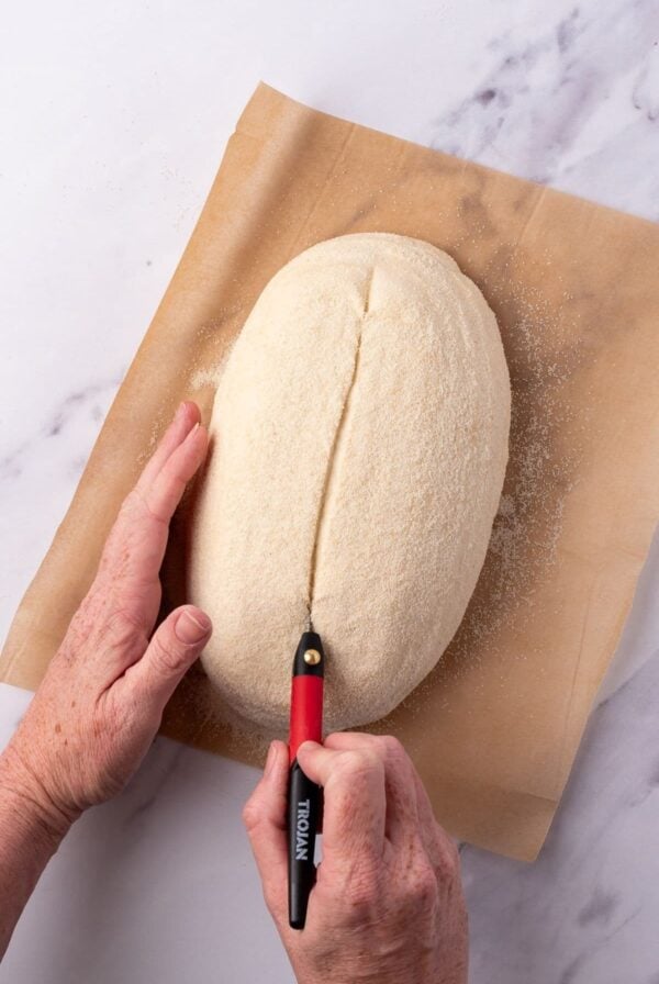 An oval loaf of sourdough (batard) being scored with a simple slash straight down the middle.